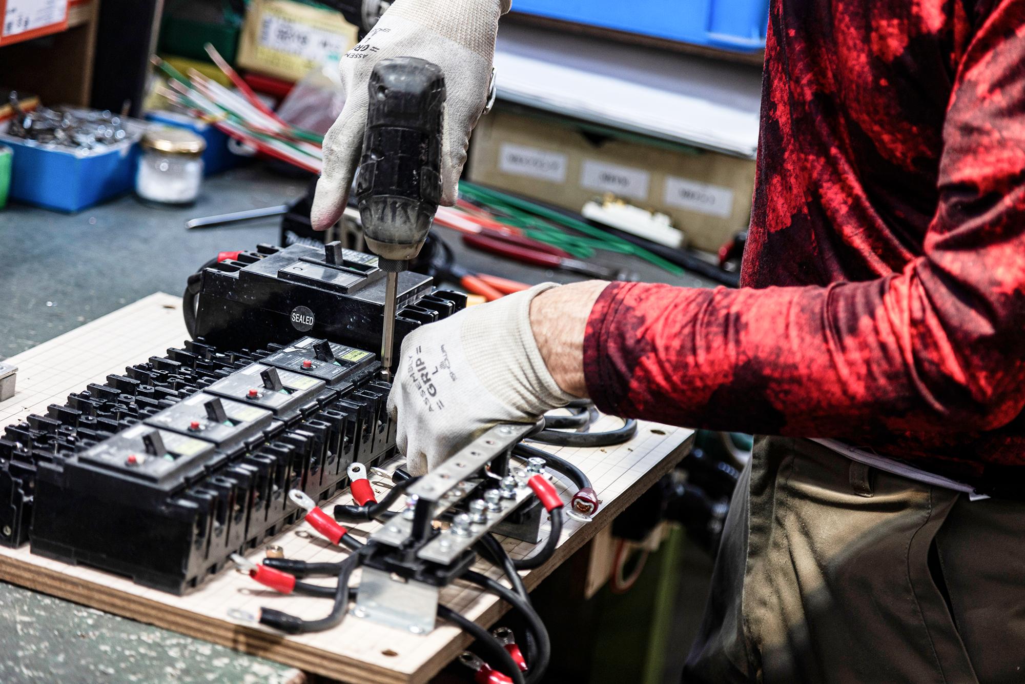 作業台上で電気部品を電動ドライバーで組み立てる技術者の手元。手袋を着用し、安全にブレーカーと配線を接続している作業工程。